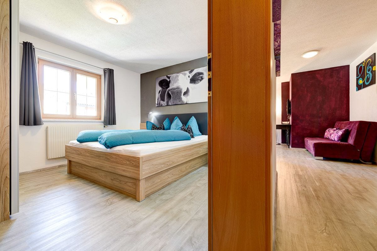 A modern bedroom with a wooden bed, a large window with curtains, a red sofa, and a wall with abstract art.