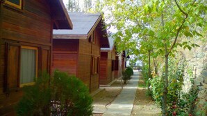 Una hilera de cabañas de madera con un estrecho sendero entre ellas, rodeadas de follaje y un muro de piedra.