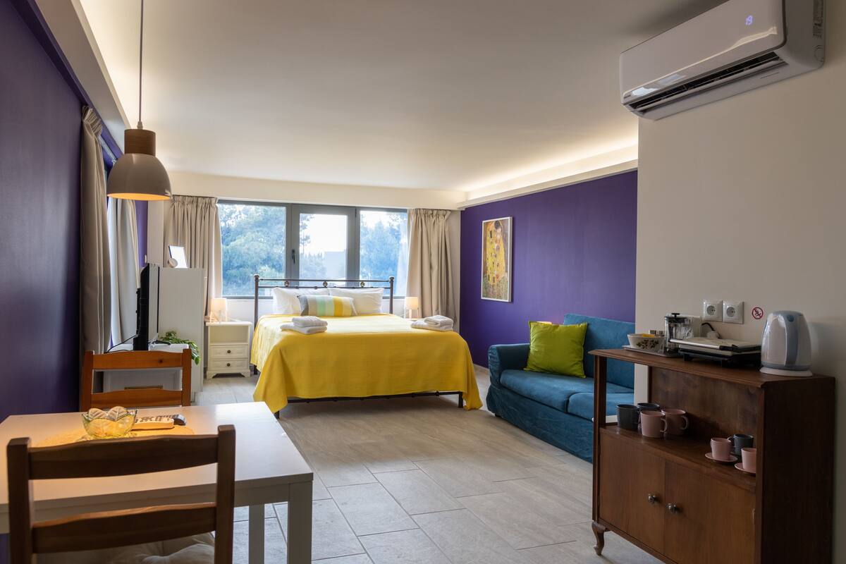 A hotel room with a bed, a sofa, a dining table, a chair, a cabinet, and an air conditioning unit.