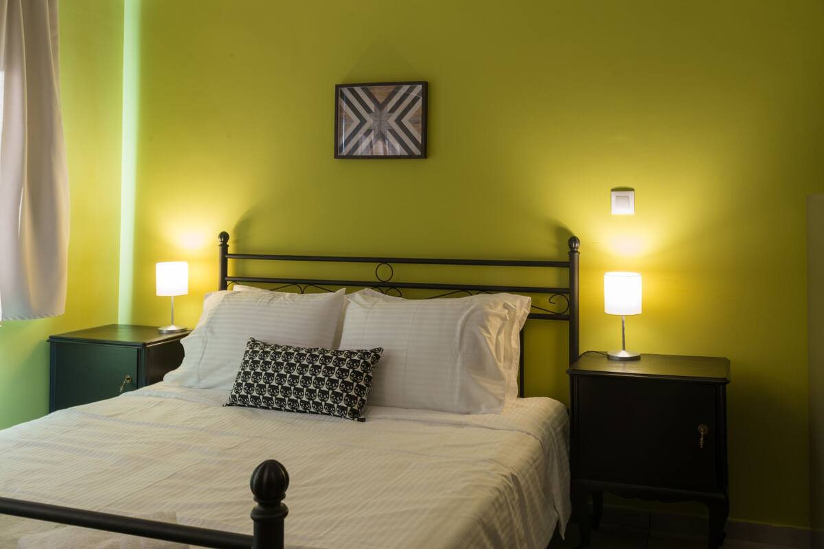 A neatly made bed with white linens and a patterned pillow, flanked by two bedside tables with lamps, in a room with green walls and a simple, modern design.