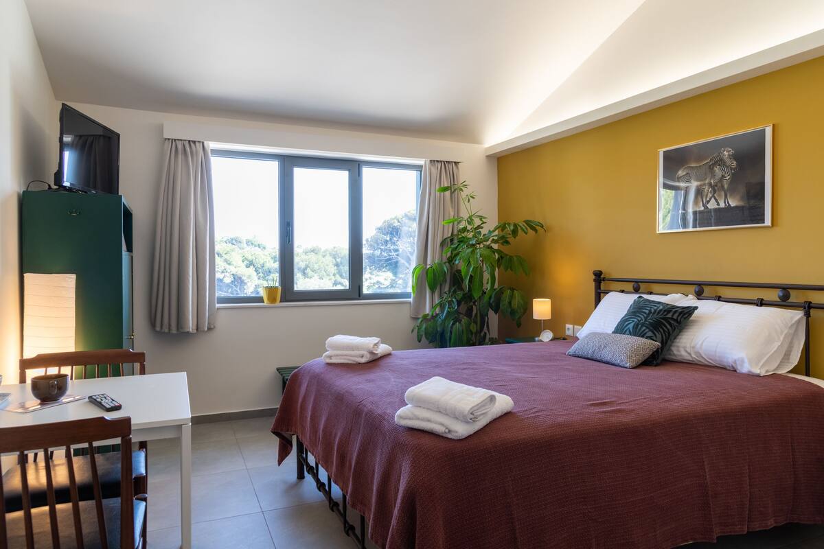 A bedroom with a bed, a desk, a chair, a window, and a plant.