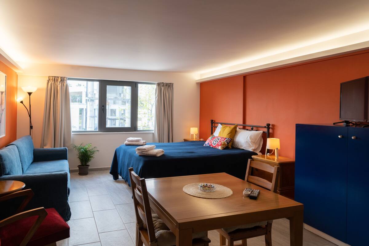 A hotel room with a bed, a desk, a chair, a sofa, a potted plant, and a TV.