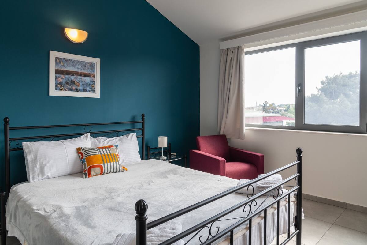 A neatly made bed with white linens and a colorful pillow, a red armchair, a nightstand with a lamp, and a painting on the wall.