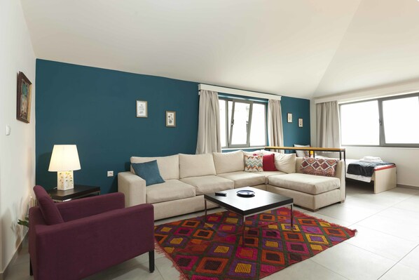Sala de estar com sofá, poltrona, mesa de centro e tapete colorido. Há uma janela com cortinas, um abajur e uma cama ao fundo.