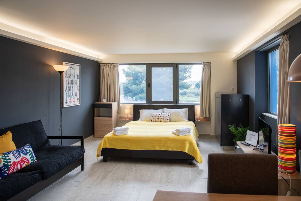 A modern hotel room with a large bed, a sofa, a desk, and a view of the outdoors.