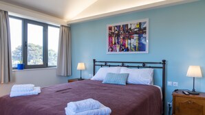 A neatly made bed with a red bedspread and two white towels, a blue abstract painting on the wall, and a window with curtains. - CHROMA Lodge Pikermi  (Rafina-Pikermi)