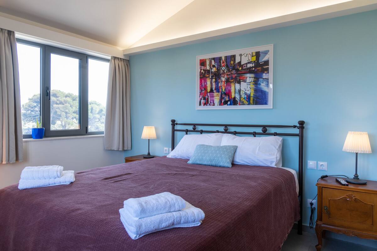 A neatly made bed with a red bedspread and two white towels, a blue abstract painting on the wall, and a window with curtains.