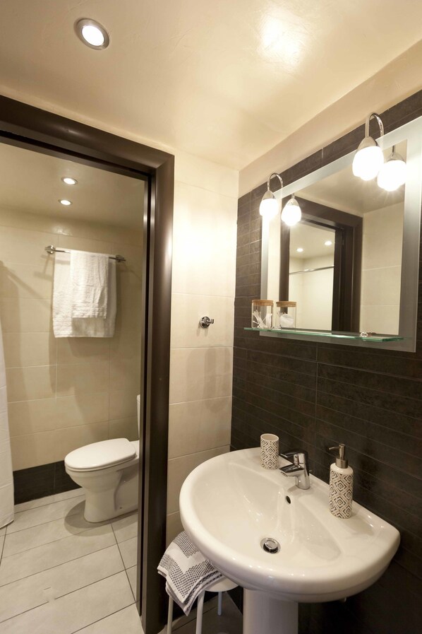 A bathroom with a white sink, dark tiled walls, a glass shower enclosure, and a white toilet. - CHROMA Lodge Pikermi (Rafina-Pikermi)