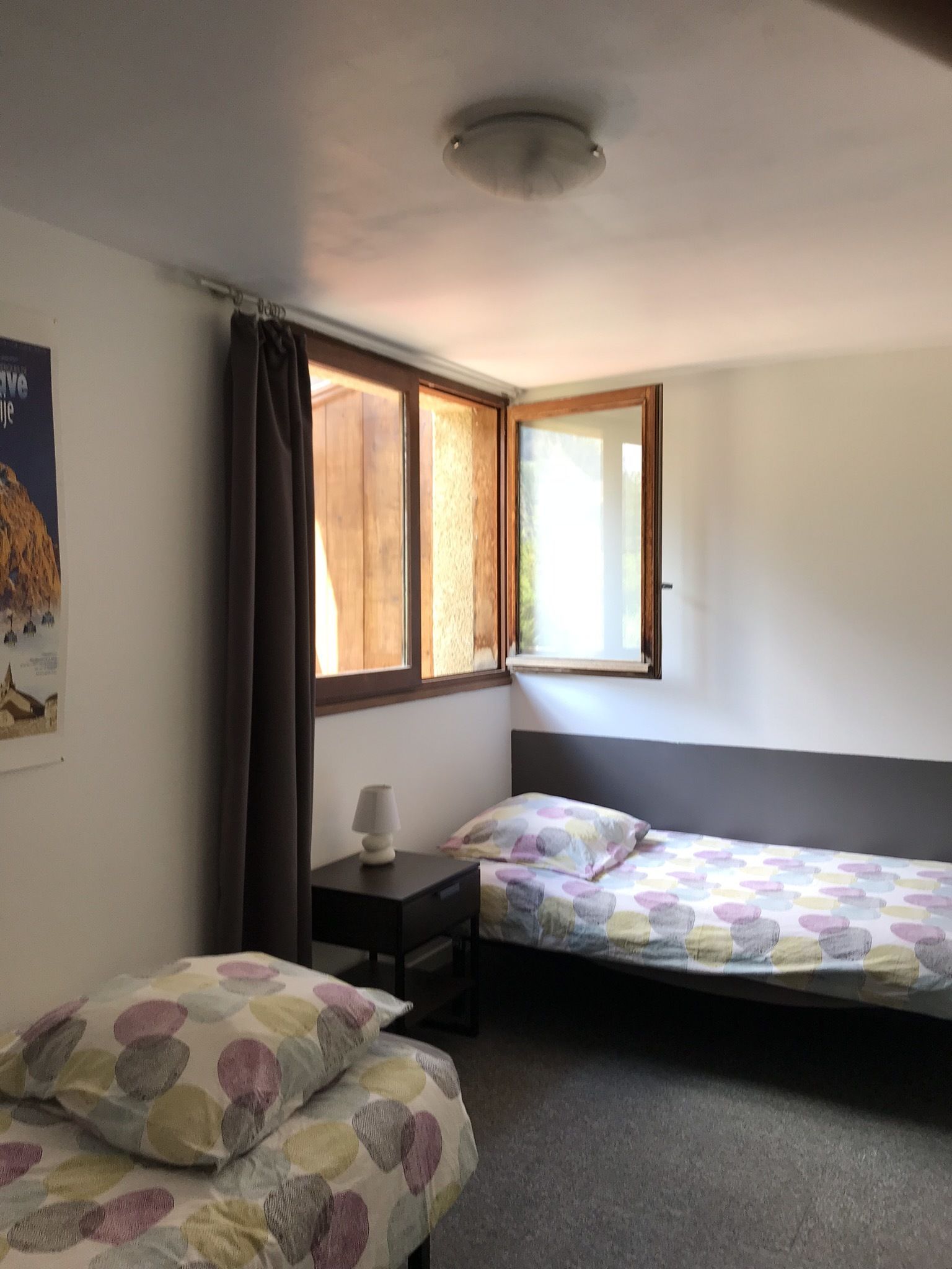 A room with two beds, a nightstand, a window with curtains, and a poster on the wall.