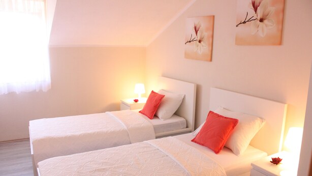 A hotel room with two beds, each with white bedding and red pillows, a nightstand with a lamp, and wall art. - Apartments Tanja & Josipa (Pag)