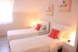 A hotel room with two beds, each with white bedding and red pillows, a nightstand with a lamp, and wall art. - Apartments Tanja & Josipa (Pag)