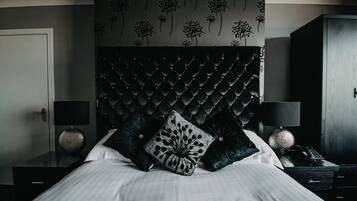 A bedroom with a tufted headboard, two black pillows, a bedside table with a lamp, and a wall with a floral pattern.
