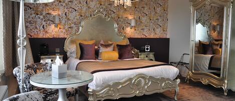A bedroom with a large ornate bed, a round glass table, a chair, and a mirror.