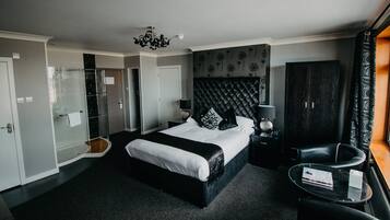 A modern hotel room with a large bed, a glass shower enclosure, a dark headboard, a black leather armchair, and a small side table with a book.