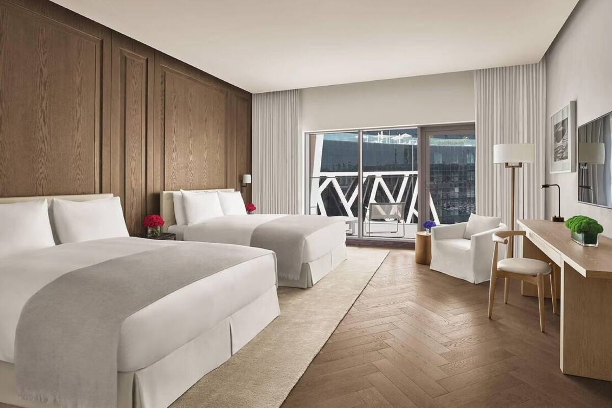 A hotel room with two beds, a desk, a chair, a balcony with a view, and a large window.