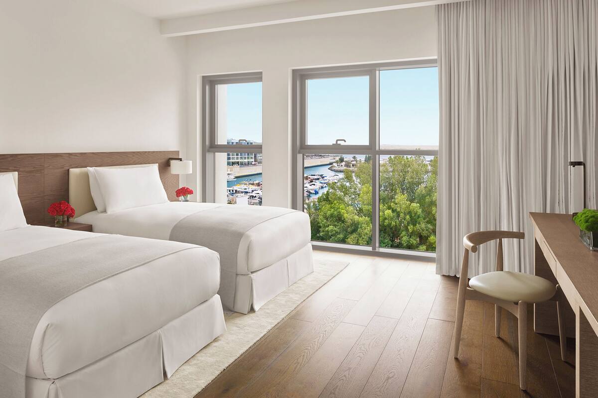 A hotel room with two beds, a desk, a chair, and large windows offering a view of a marina and cityscape.