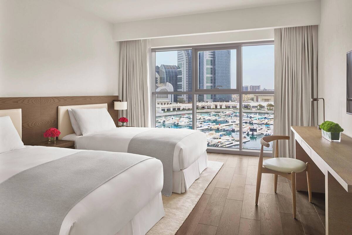 A hotel room with two beds, a desk, a chair, a TV, and a large window with a city view.