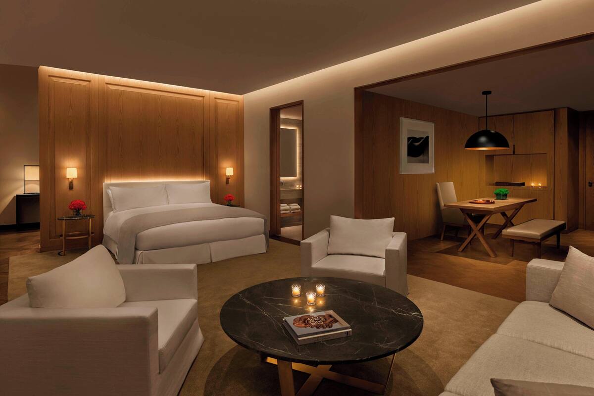 A modern hotel room with a large bed, two white armchairs, a round coffee table, and a small dining area with a table and chairs.