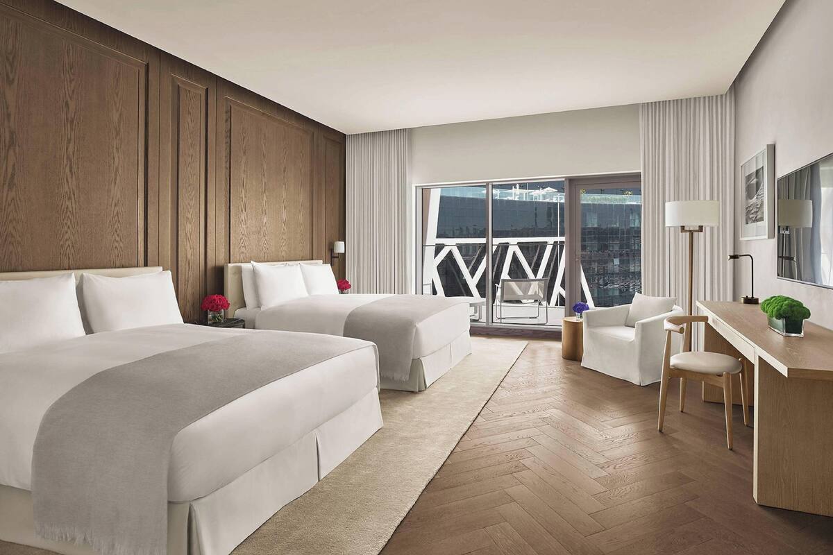 A hotel room with two beds, a desk, a chair, and a balcony with a city view.