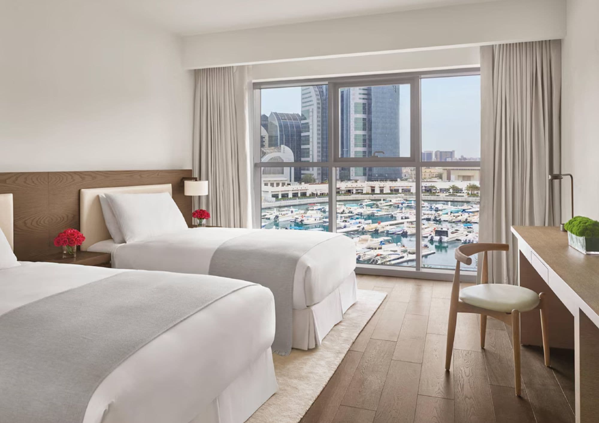 A hotel room with two beds, a desk, a chair, and a large window with a marina view.