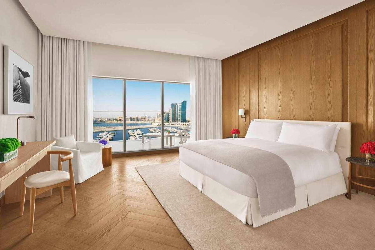 A modern hotel room with a large bed, a desk, a chair, and a view of a cityscape through large windows.