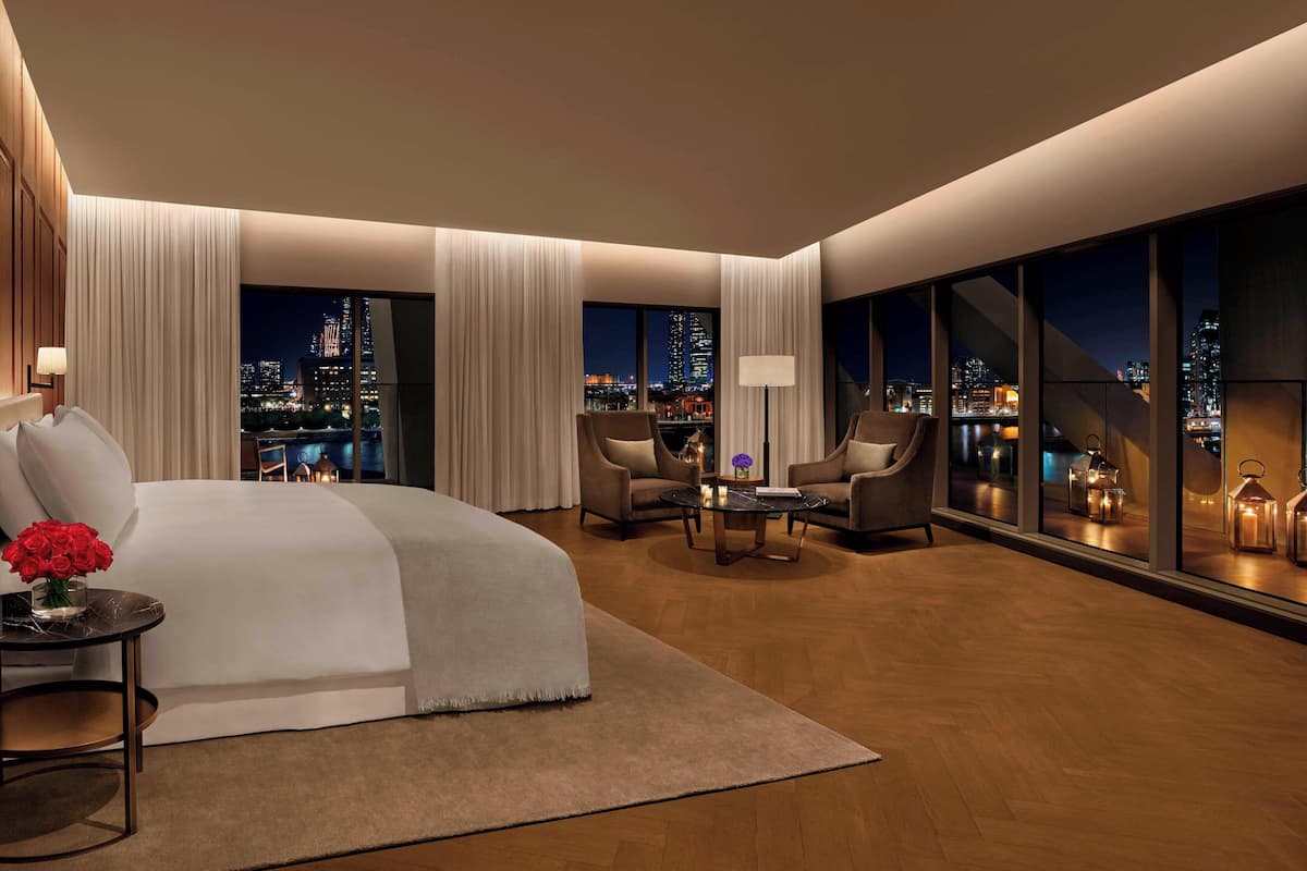 A modern hotel room with a large bed, two armchairs, a coffee table, and a view of the cityscape at night.