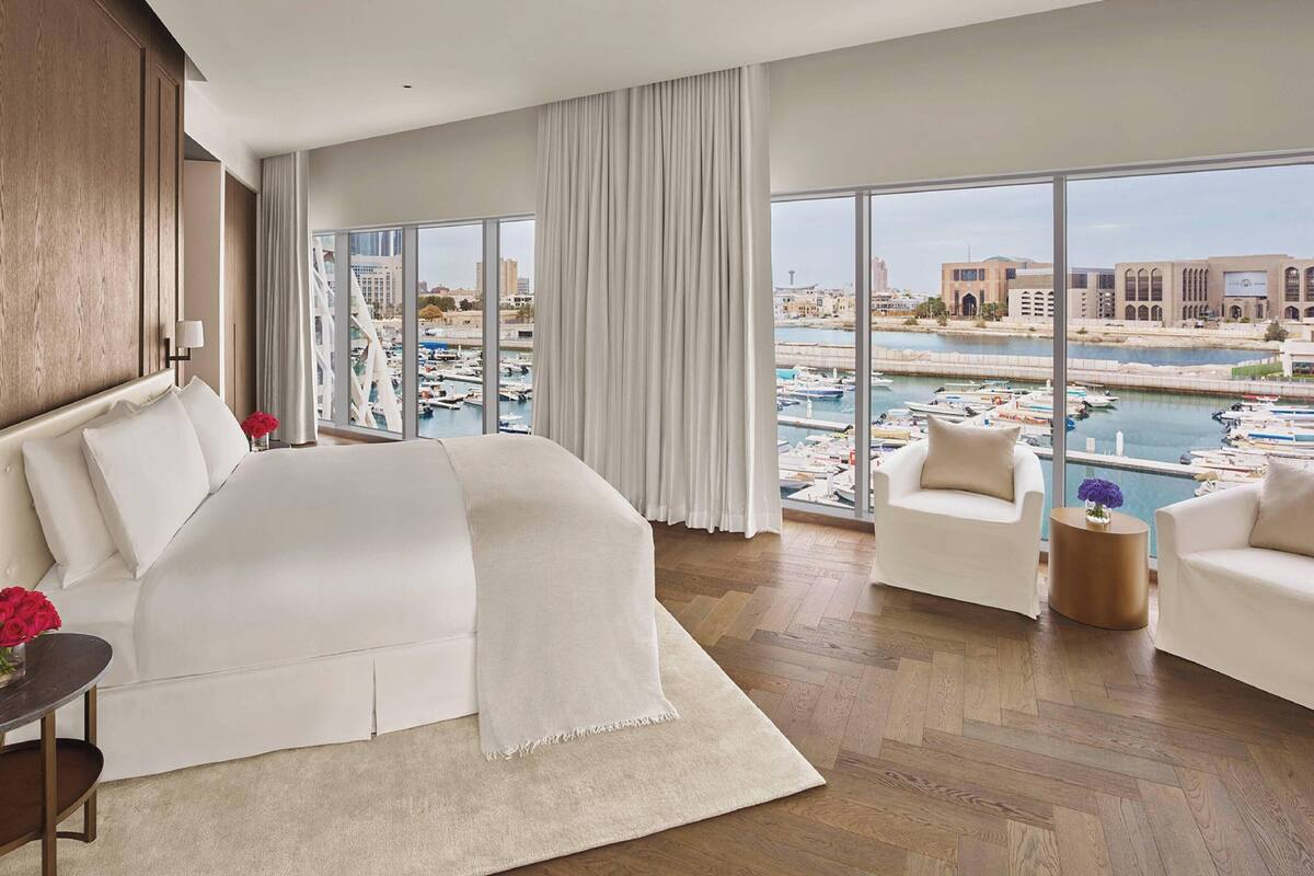 A spacious hotel room with a large bed, two armchairs, a wooden floor, and a view of a marina with boats.