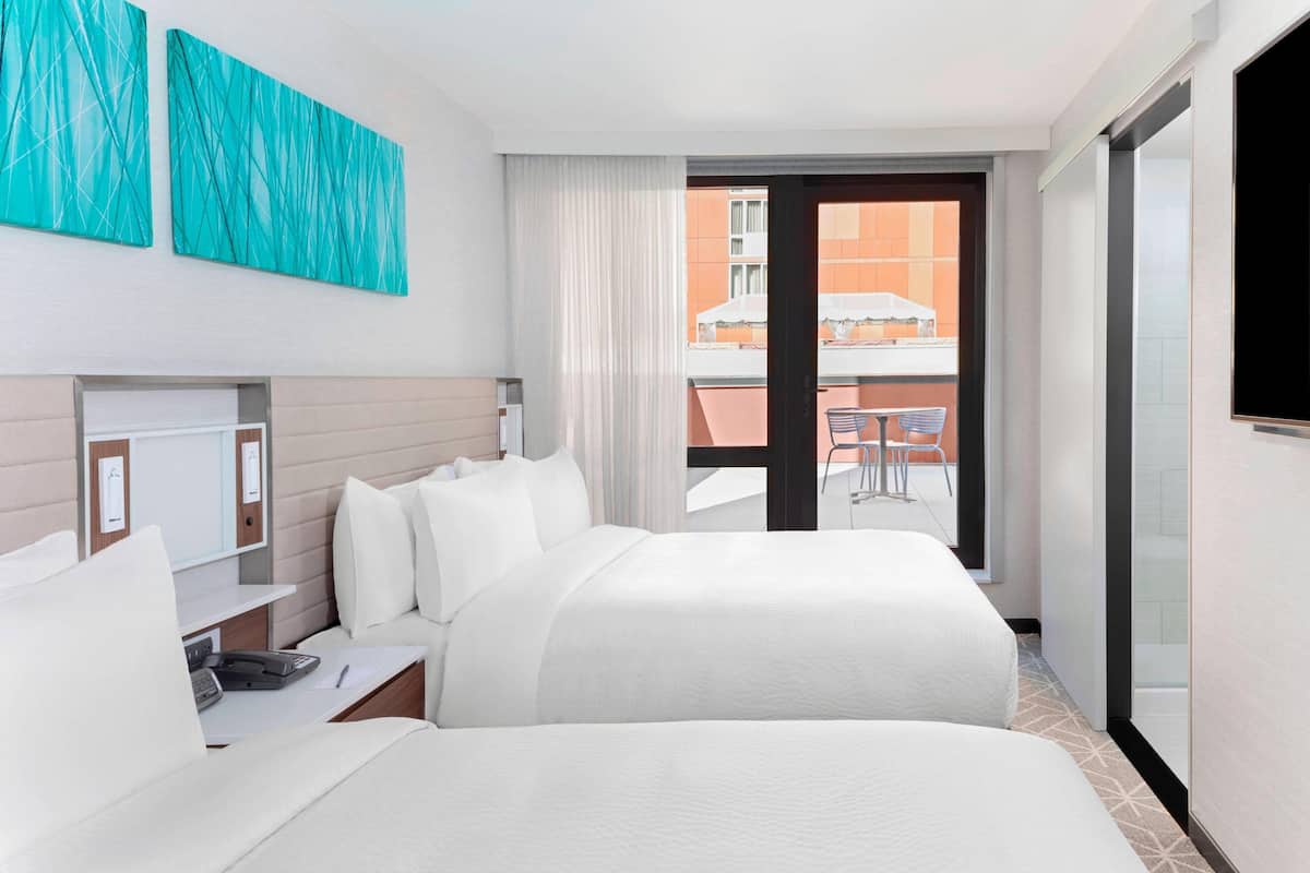 Une chambre d’hôtel avec deux lits, un bureau, une chaise, une télévision et un balcon donnant sur des immeubles.