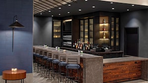 Zona de bar moderna con una barra alargada, varios taburetes y un estante en la pared con botellas.