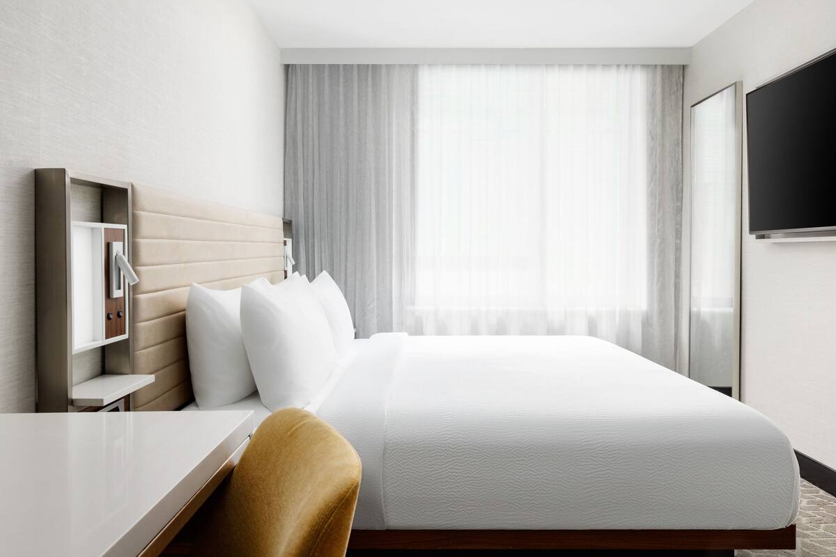 Une chambre d’hôtel avec un lit, un bureau, une chaise, une télévision et une fenêtre avec des rideaux.