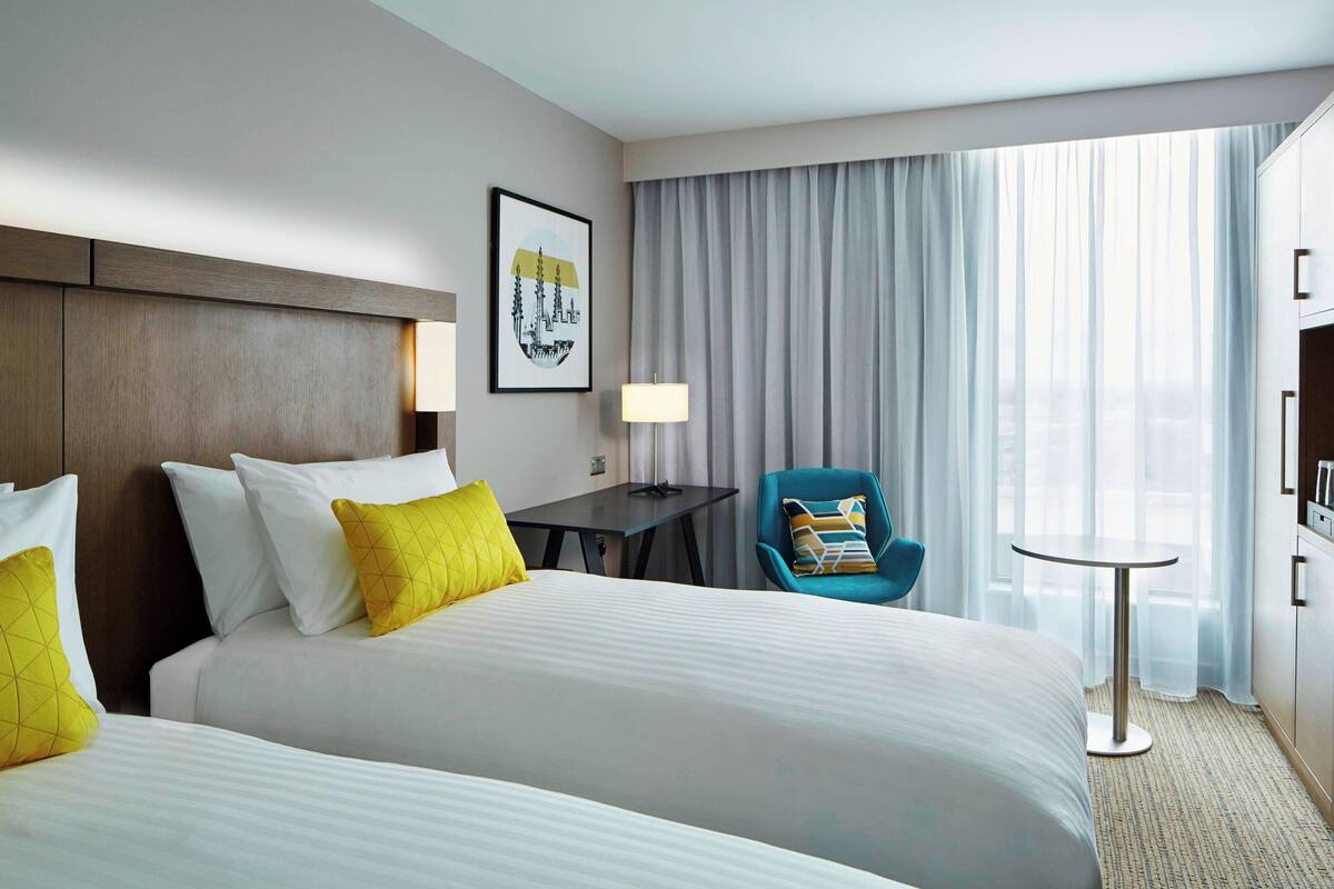 Habitación de hotel con dos camas, un escritorio, una silla, una lámpara y vistas a la ciudad a través de amplias ventanas.