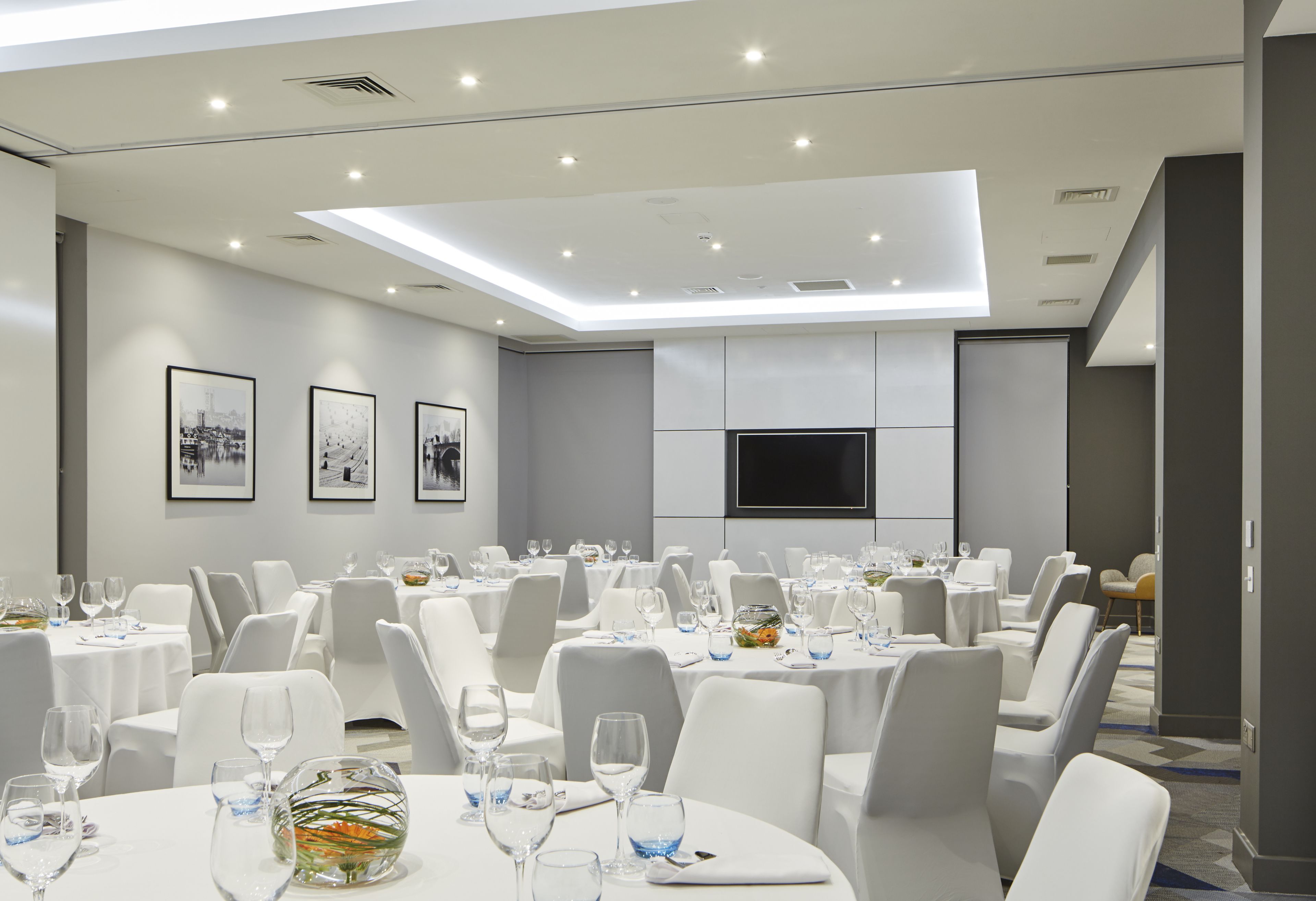 A conference room with multiple tables, white chairs, and a flat-screen TV on the wall.