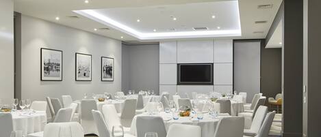 A conference room with multiple tables, white chairs, and a flat-screen TV on the wall.