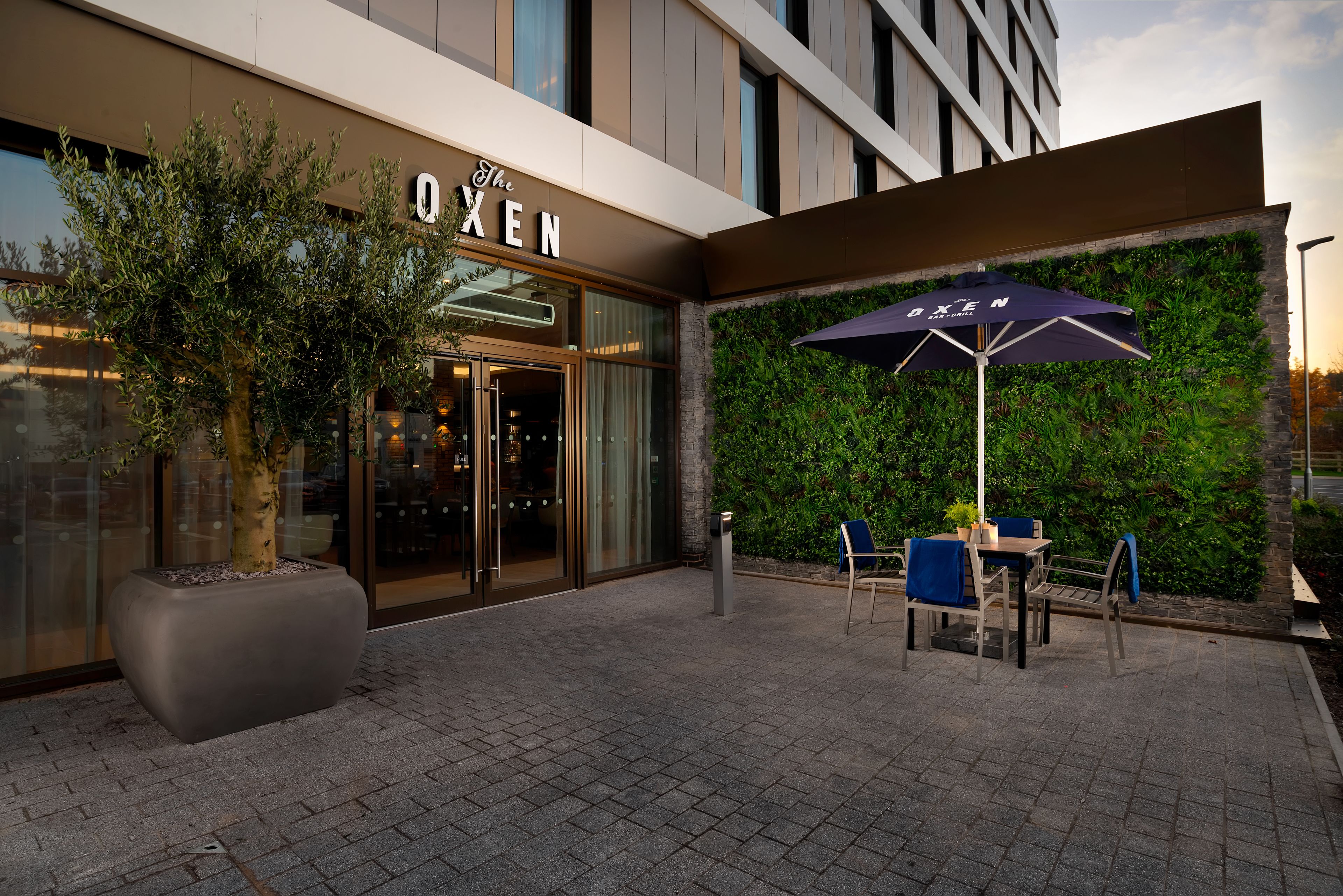 A hotel entrance with a glass door, outdoor seating area with a table and chairs, and a potted plant.
