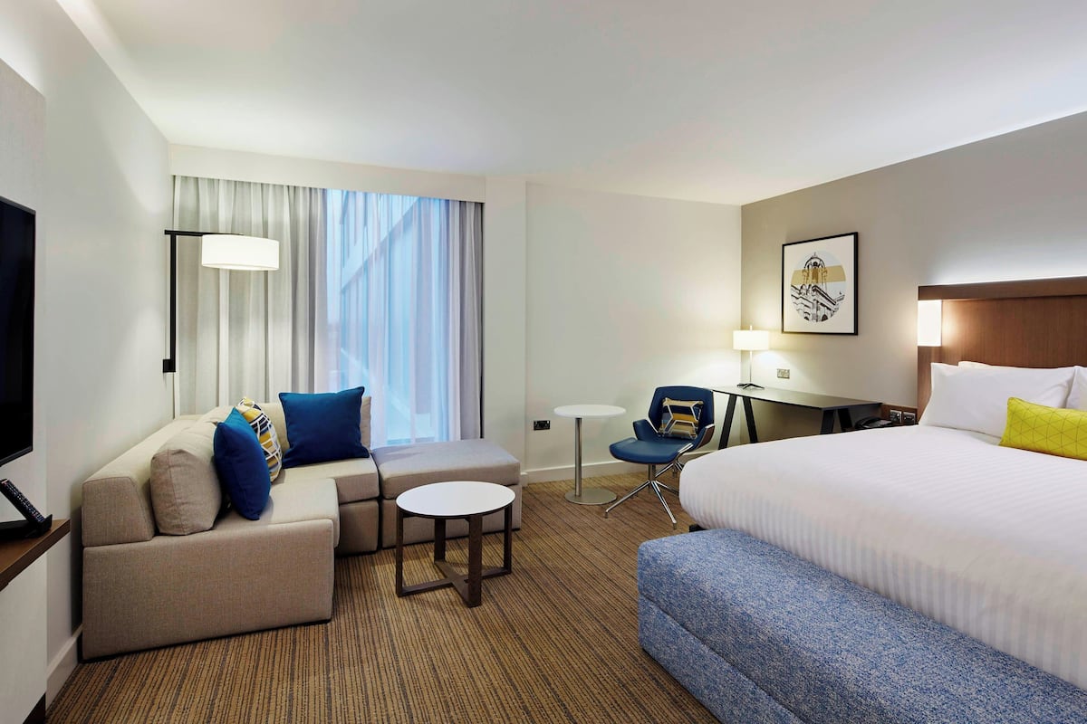 Habitación de hotel con una cama grande, un sofá, una mesita pequeña y una silla. Hay un televisor a la izquierda y un escritorio con una lámpara a la derecha.