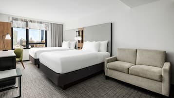 Habitación de hotel con dos camas, un sofá, un escritorio y un amplio ventanal con vistas a la ciudad.