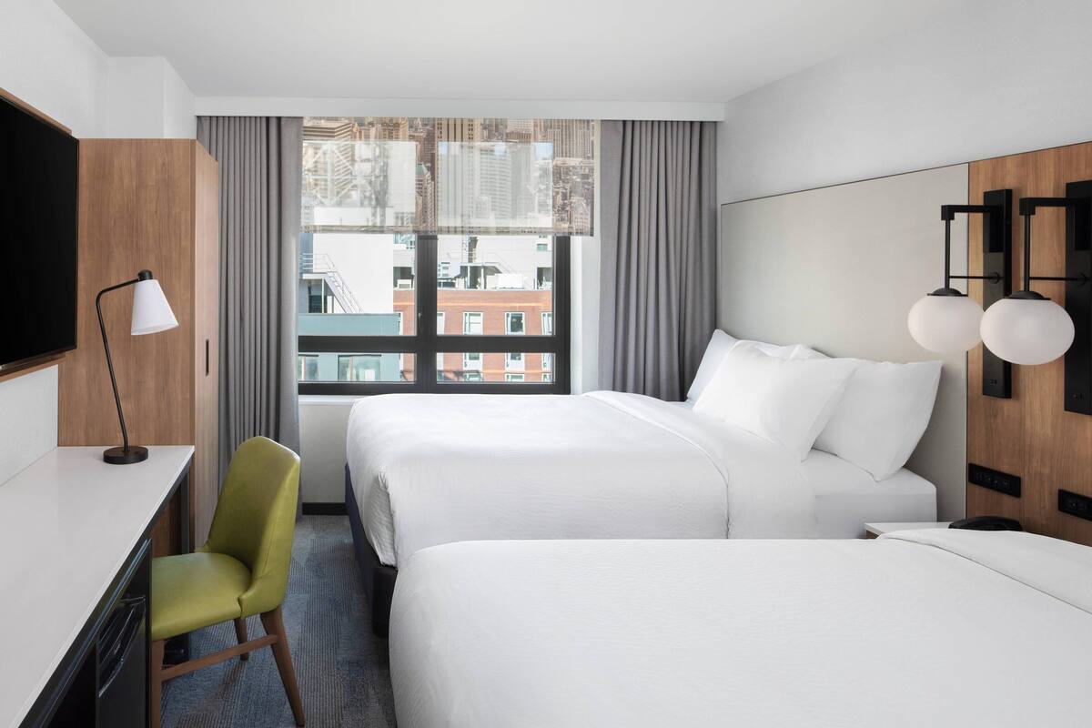 Une chambre d’hôtel avec deux lits, un bureau, une chaise, une télévision et une fenêtre donnant sur la ville.