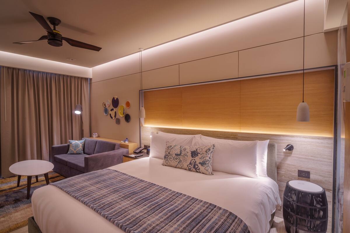 A modern hotel room with a large bed, a sofa, a small table, and a ceiling fan.