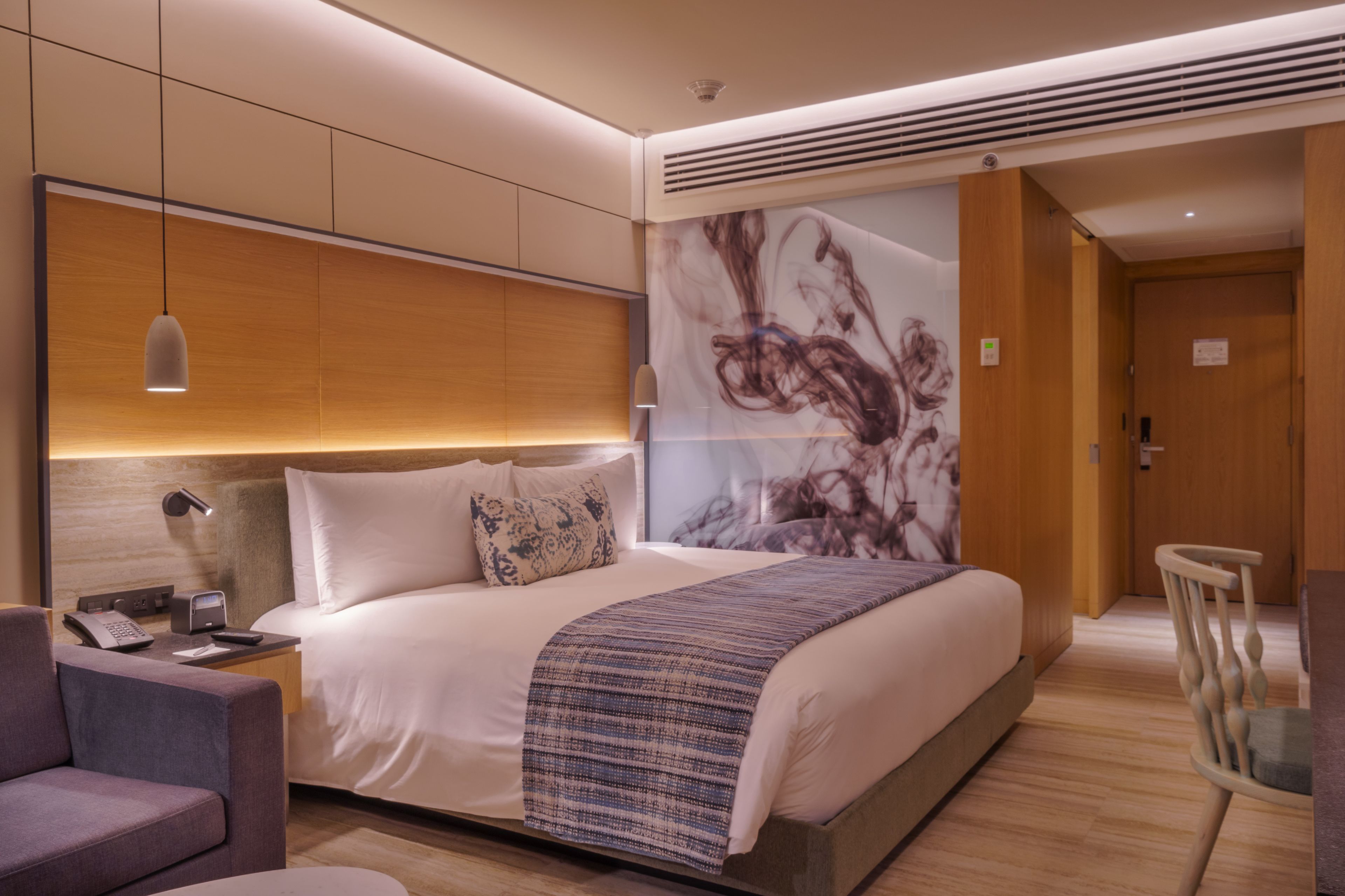 Habitación de hotel moderna con una cama grande, mesita de noche con lámpara, una silla y un cuadro abstracto de gran tamaño en la pared.