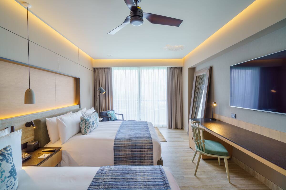 A hotel room with two beds, a desk, a chair, a television, and a ceiling fan.