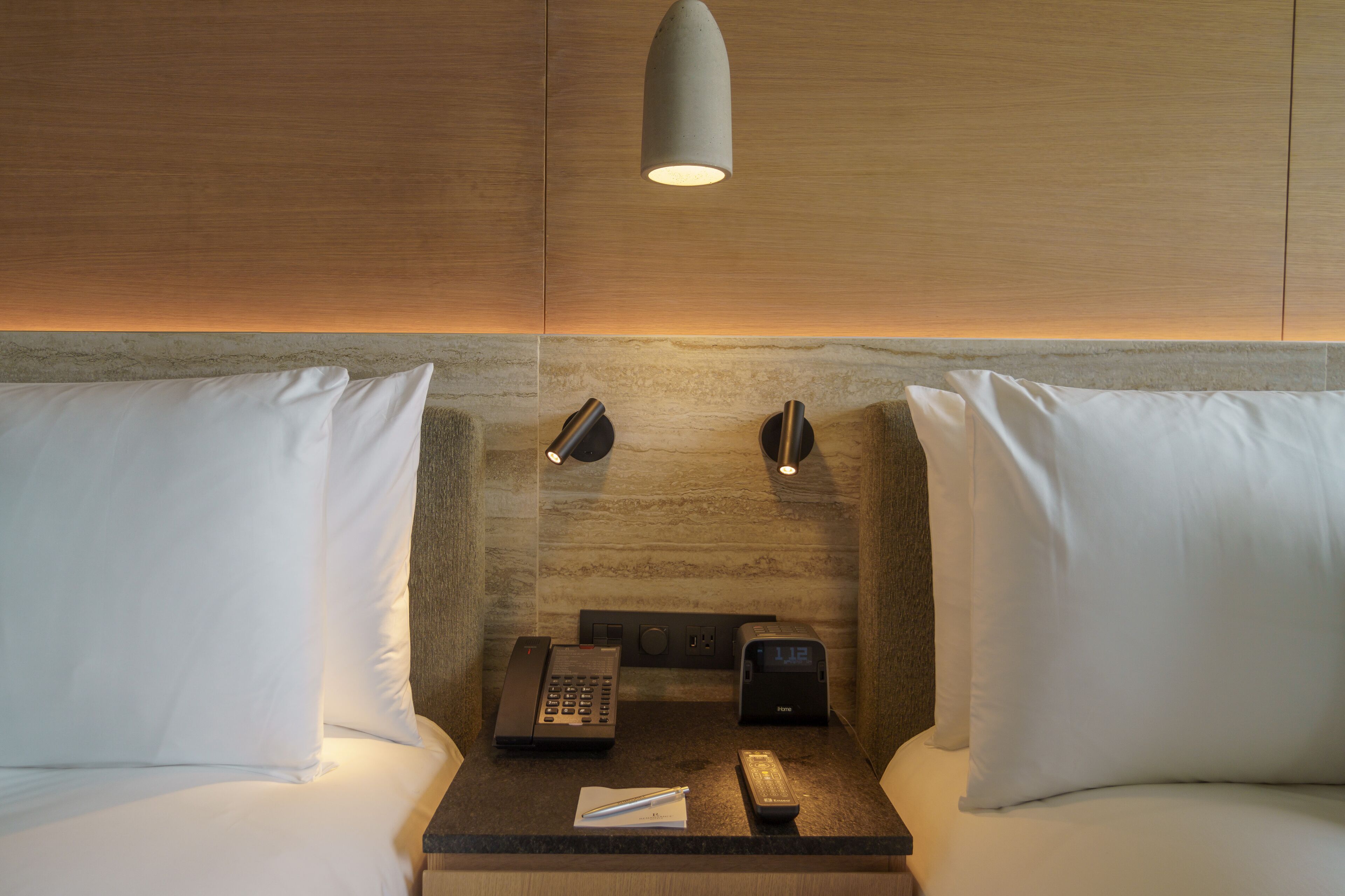 A hotel room with a wooden headboard, two bedside lamps, a telephone, a digital clock, and a nightstand with a pen and remote control.