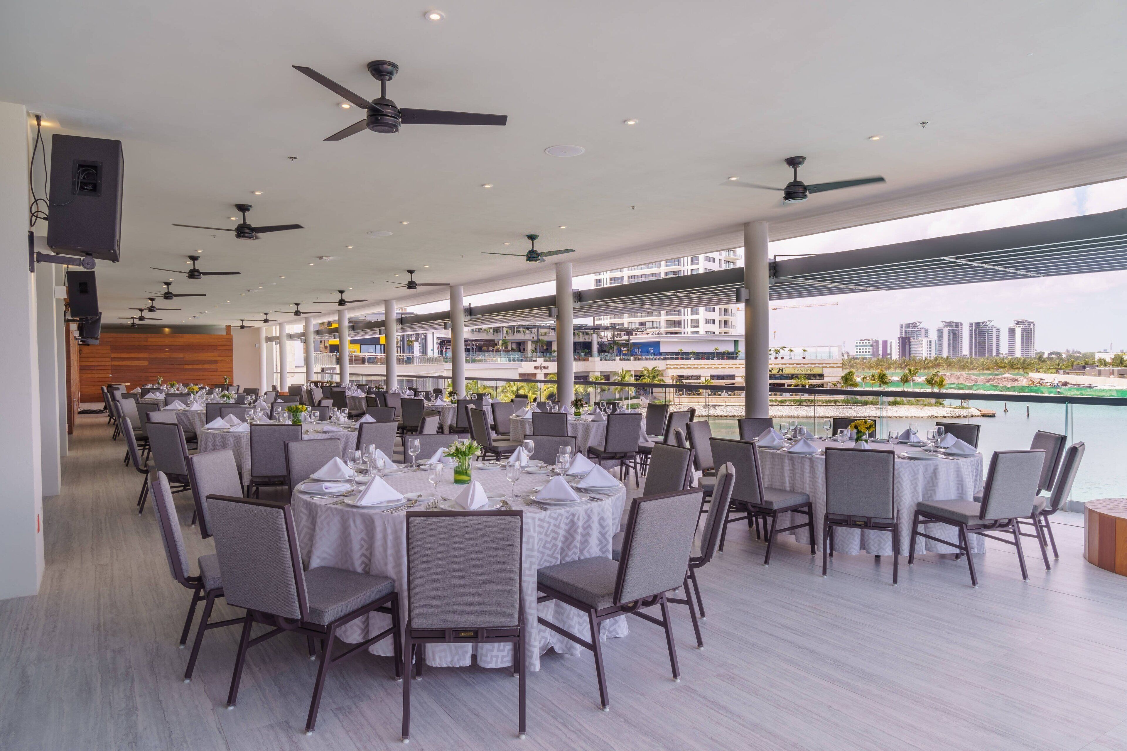 Un vaste espace extérieur aménagé pour les repas, avec plusieurs tables et chaises rondes, des ventilateurs de plafond et une vue sur les bâtiments d’une ville et sur un plan d’eau.