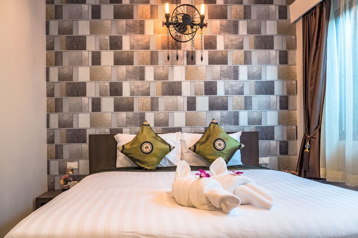 A hotel room with a tiled wall, a chandelier, a bed with white bedding, and two decorative pillows.
