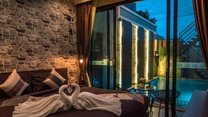 Ein Schlafzimmer mit Steinwand, einem Bett mit einem schwanenformigen Handtuch, Blick auf einen Swimmingpool und einer Treppe.