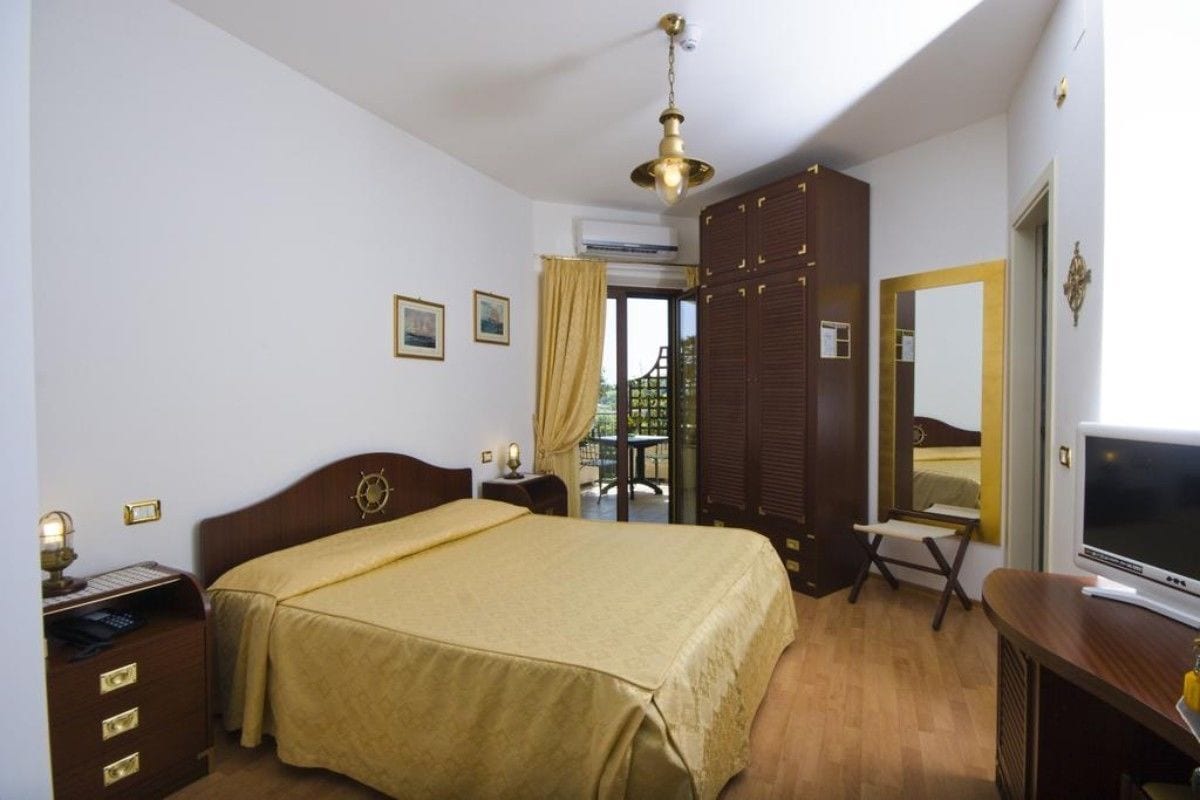 A hotel room with a bed, a desk with a computer, a TV, a wardrobe, and a window with curtains.