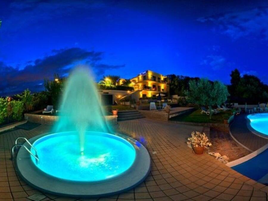 A nighttime scene of a swimming pool area with a fountain, surrounded by a landscaped garden and a building with illuminated windows.
