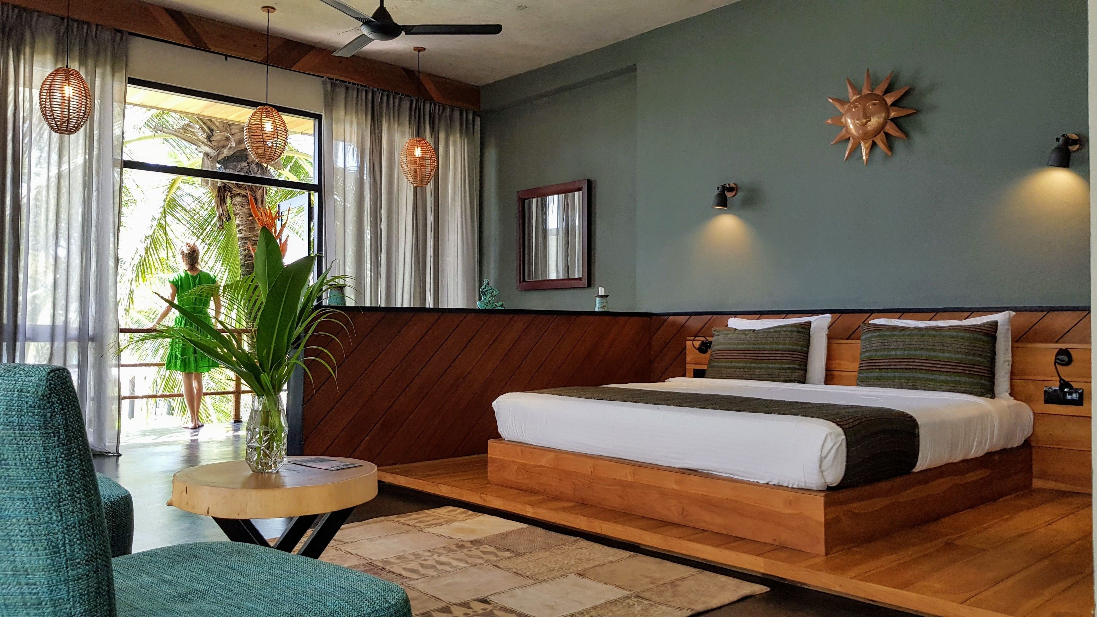 A bedroom with a wooden bed, a small round table, a green chair, and a large window with curtains.