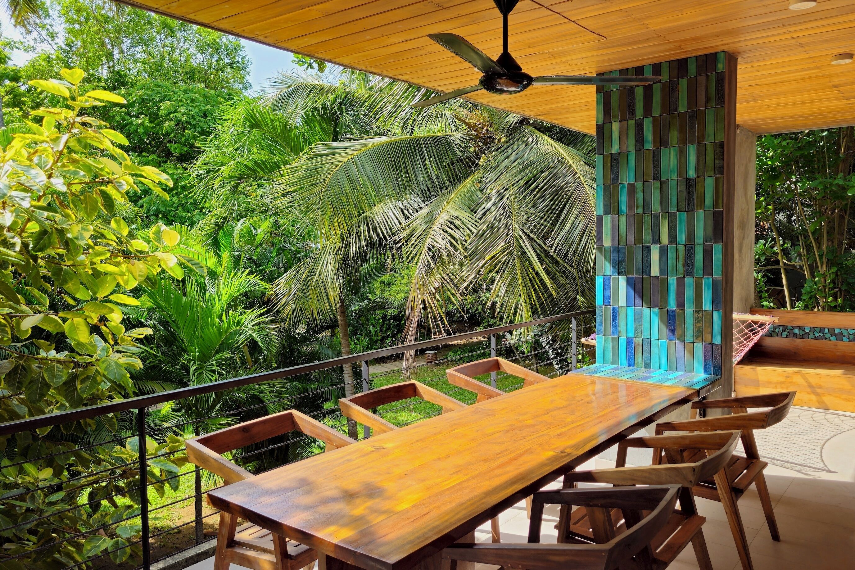 An outdoor dining area with a wooden table and chairs, surrounded by lush greenery and a decorative wall.