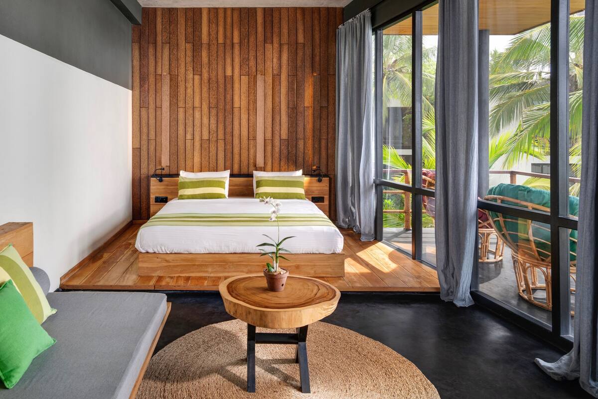 A modern bedroom with a wooden headboard, a large bed with green and white bedding, a round wooden table with a potted plant, and a view of a balcony with wicker furniture and a palm tree.