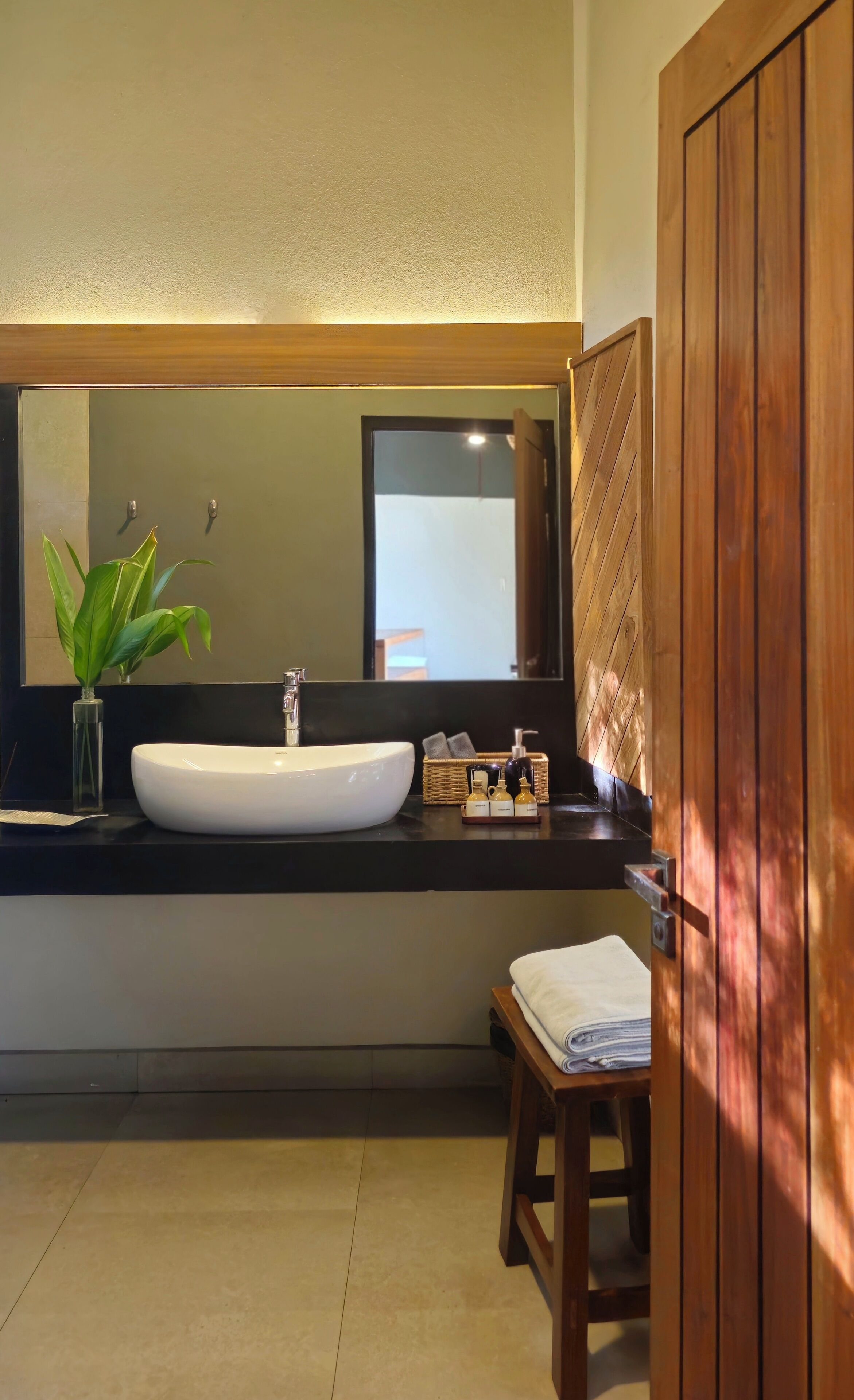 A modern bathroom with a large mirror, a white sink, a small plant, and a wooden door.